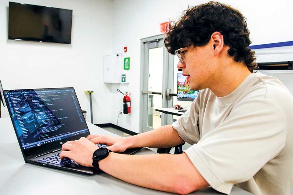 Person seated at a desk in a workshop or lab environment, working on a mechanical or engineering project. The desk has various tools and components, including wires, metal parts, and electronic equipment. The background shows shelves with additional suppl