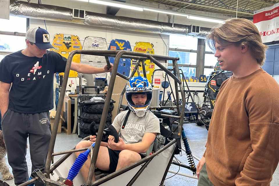 A person wearing a helmet rides a bicycle on a paved path surrounded by autumn trees with colorful leaves. The cyclist is in motion, and the background shows a mix of green and orange foliage, suggesting a fall season setting.
